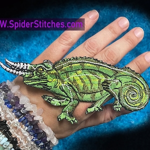 Könnte beinhalten: Grüner gestickter Aufnäher eines Chamäleons mit weißen Hörnern und einem eingerollten Schwanz. Der Aufnäher befindet sich auf einer Hand mit einem Stapel Armbänder.