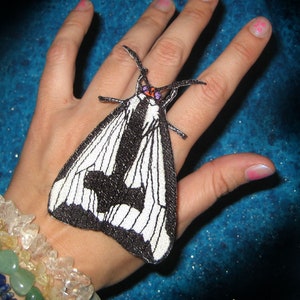 May include: A black and white moth with a white stripe down the center of its wings. The moth is on a person's hand, which is wearing a bracelet with colorful stones.