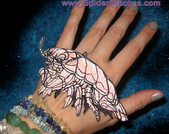 May include: A pink and black embroidered patch of a sea creature with segmented legs and a long antenna. The patch is on a person's hand.