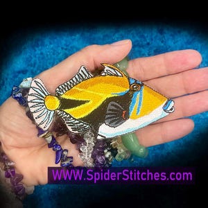 May include: Embroidered patch of a tropical fish with yellow, black, and blue details. The fish has a white and black tail fin. The patch is held in a hand, with beaded bracelets in shades of purple and blue. Text on the image reads "WWW.SpiderStitches.com".