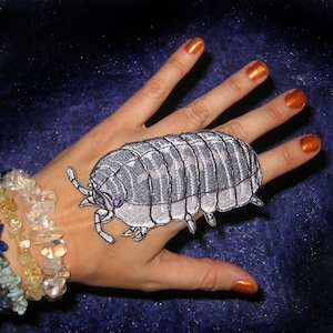May include: A hand model wearing a silver and black embroidered patch of a pill bug on the back of their hand.