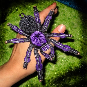 May include: A hand holding a purple and black embroidered tarantula patch. The patch has a detailed design with a purple abdomen and black legs.