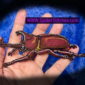 May include: Embroidered patch of a rhinoceros beetle in shades of burgundy, brown, and yellow. The insect has detailed legs and antennae. The text "WWW.SpiderStitches.com" is displayed on a purple banner.