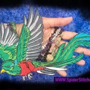 May include: Embroidered patch of a vibrant Quetzal bird with green, red, and blue details. The bird has outstretched wings and long tail feathers. The patch is held against a blue background. Website text: WWW.SpiderStitches.com.