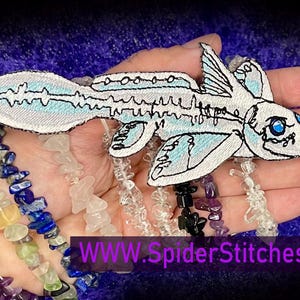 Può includere: Un toppa ricamata bianca che rappresenta un pesce stilizzato con occhi blu. Il pesce ha un corpo lungo e sottile e una coda grande a forma di ventaglio. La toppa è su una mano con una varietà di braccialetti di pietre preziose colorate. Il testo "WWW.SpiderStitches.com" è visibile nell'angolo in basso a destra dell'immagine.