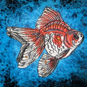 May include: A red and white goldfish with black outlines is embroidered on a blue and white speckled background. The fish is swimming to the right with its fins spread out.