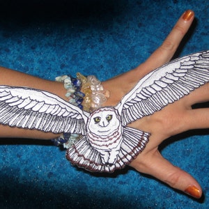 Op de afbeelding: Een witte en zwarte geborduurde uil met een bruin oog, die op de arm van een persoon zit. De uil draagt een kralenarmband met gekleurde stenen.