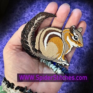May include: An embroidered patch depicting a chipmunk with a long, bushy tail. The patch is brown and white and is being held in a hand wearing a bracelet made of stones. The text "WWW.SpiderStitches.com" is visible at the bottom of the image.