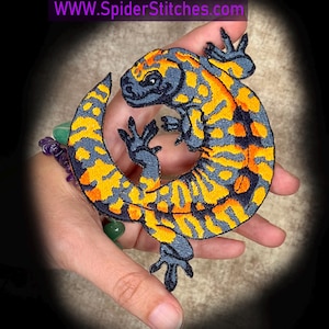 Könnte beinhalten: Gestickter Aufnäher mit einem Salamander-Design. Der Salamander hat ein detailliertes Muster aus gelben und orangefarbenen Flecken auf einem dunkelgrauen Hintergrund. Der Aufnäher wird in einer Hand gehalten, mit der Website-Adresse WWW.SpiderStitches.com oben.
