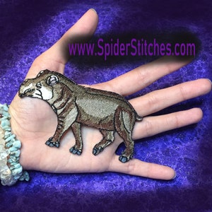 Puede incluir: Parche bordado para planchar de un tapir marrón con el hocico blanco, de pie sobre sus cuatro patas. El parche está en una mano, con el sitio web www.spiderstitches.com visible en el fondo.