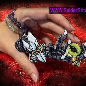 May include: Black and white embroidered patch of a clawed hand with a large green eye. The hand is holding a crystal with gold accents. The patch is on a red background. WWW.SpiderStitches.com