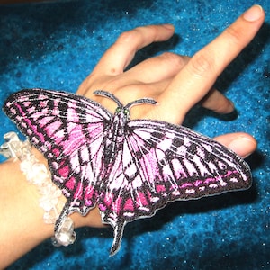 May include: A pink and black butterfly embroidered patch with white accents. The patch is attached to a person's wrist.