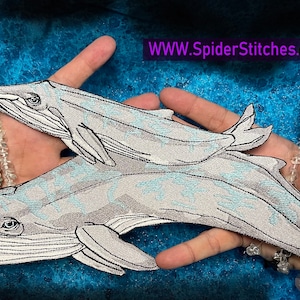 Può includere: Un paio di toppe ricamate a forma di balena grigia, una più grande dell'altra, con la balena più piccola che nuota accanto a quella più grande. Le toppe sono mostrate su una mano con diversi braccialetti.