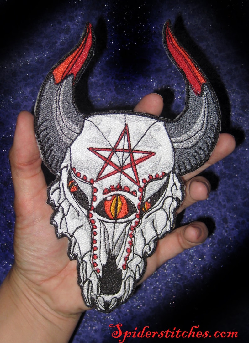 May include: An embroidered patch featuring a white skull with a red pentagram and orange eyes. The skull has black horns and a detailed design. The patch is held in a hand against a blue background.