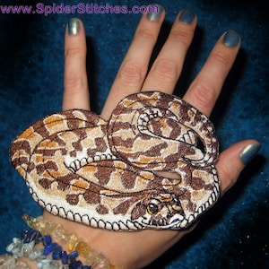 Puede incluir: Un parche bordado que representa una serpiente enroscada con una cabeza y escamas detalladas. El parche es marrón y blanco y se muestra en la mano de una persona.