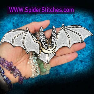 May include: An embroidered patch shaped like a bat with light gray wings and a tan body, featuring black stitched outlines and details. The text WWW.SpiderStitches.com is visible at the top of the image in a purple font against a dark background.