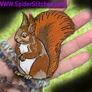 Puede incluir: Parche bordado de una ardilla marrón con cola tupida, sosteniendo una nuez en sus patas. El parche está en una mano, con un fondo verde.