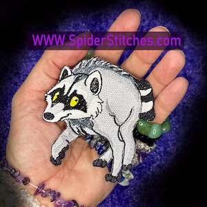 May include: An embroidered patch featuring a stylized raccoon with light gray fur, black eye markings, and bright yellow eyes. The patch includes a striped tail and black paws. The text WWW.SpiderStitches.com is visible across the top of the image.