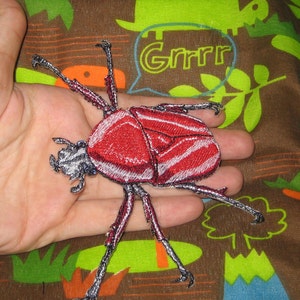 Peut inclure: Patch brodé représentant un scarabée rouge et blanc avec des pattes noires et des accents argentés. Le patch est sur un fond de tissu brun avec un motif vert et bleu.