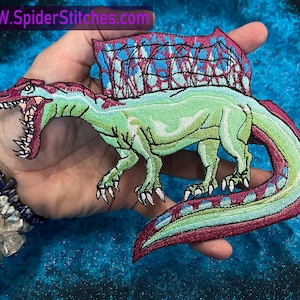 May include: Embroidered patch of a green Spinosaurus dinosaur with a red and blue sail on its back. The dinosaur is depicted with an open mouth and sharp teeth. The patch is held in a hand.
