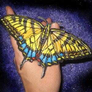 Op de afbeelding: Een geborduurde vlinderpatch in geel en zwart met blauwe en rode accenten. De patch bevindt zich op een hand tegen een donkerblauwe achtergrond.