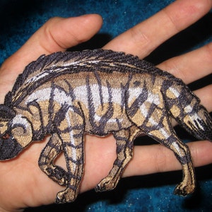 May include: Embroidered patch of a brown and black African wild dog, also known as a painted dog, with white stripes. The patch is on a hand.