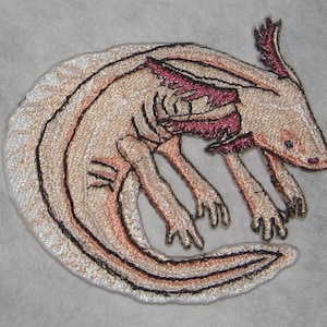 May include: Embroidered patch of an axolotl, a type of salamander, with a pink and white color scheme. The axolotl is curled up with its tail tucked under its body.