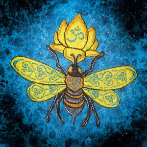 May include: An embroidered bee with yellow wings and a lotus flower on its head. The bee has intricate details and the lotus flower has an Om symbol in the centre. The background is a blue and black galaxy-like pattern.
