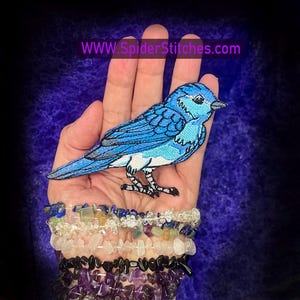 May include: Bluebird embroidered patch with white accents. The patch is shown on a hand with a stack of bracelets.