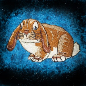 Può includere: Un coniglio marrone e bianco con grandi orecchie cadenti è ricamato su uno sfondo blu con una texture scintillante.