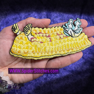 May include: Yellow embroidered iron-on patch of a sea slug with a blue and white flower on top. The patch is on a hand. The text "WWW.SpiderStitches.com" is visible below the hand.