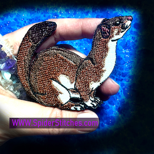 Puede incluir: Un parche bordado de una comadreja o armiño con la cola enroscada. El parche es marrón y blanco y está en una mano con un fondo azul.