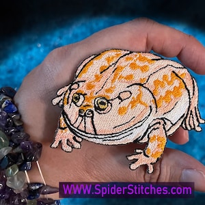 Pode incluir: Um remendo bordado de um sapo laranja claro e branco com olhos pretos. O sapo está sentado em uma mão com uma pulseira no pulso. O texto "WWW.SpiderStitches.com" está na parte inferior da imagem.