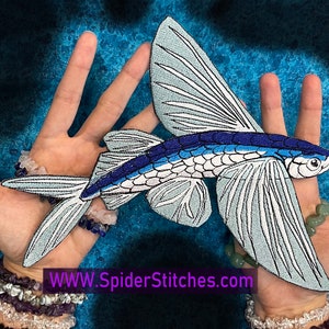 Könnte beinhalten: Ein detaillierter, gestickter Aufnäher eines fliegenden Fisches, mit Blau-, Weiß- und Schwarztönen. Der Aufnäher hat ausgebreitete Flügel und einen stromlinienförmigen Körper. Der Text "WWW.SpiderStitches.com" ist sichtbar. Der Hintergrund ist strukturiert blau.