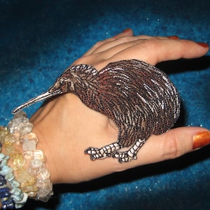 May include: A brown and white kiwi bird patch with a long beak and detailed feathering. The patch is an embroidered design, perfect for adding to clothing or accessories. The bird is shown on a hand.