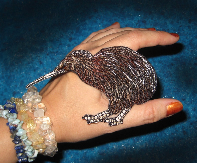 Op de afbeelding: Een geborduurd kiwi vogel patch, bruin en wit met een lange snavel en gedetailleerde veren. Perfect om toe te voegen aan kleding of accessoires. De vogel wordt op een hand getoond.