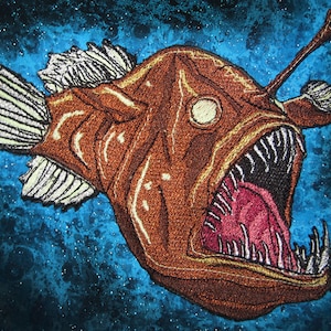May include: Embroidered patch featuring a brown anglerfish with an open mouth, visible teeth, and a glowing lure. Set against a dark blue background with white speckles, reminiscent of a starry sky or the deep sea.