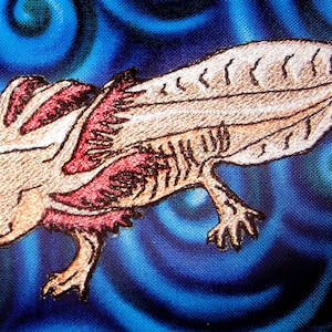 May include: Embroidered axolotl patch with a light brown body, red gills, and black outlines. The axolotl is on a blue background with swirling patterns.