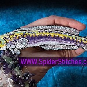 Può includere: Una mano tiene un toppa ricamata a forma di pesce bianco, viola e giallo. La toppa è lunga circa 12,7 cm e larga 5 cm. Il testo "WWW.SpiderStitches.com" è visibile sotto la toppa.