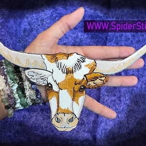 May include: A white and brown embroidered patch of a longhorn steer head with large horns. The patch is held in a hand against a purple background. The text "WWW.SpiderStitches.com" is visible in the top right corner of the image.