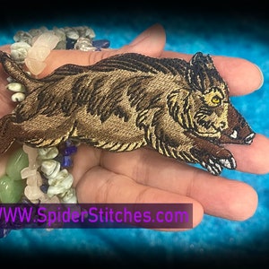 May include: A brown and gold embroidered patch of a wild boar with a yellow eye. The patch is on a hand with a bracelet of colorful stones. The text "WWW.SpiderStitches.com" is at the bottom of the image.