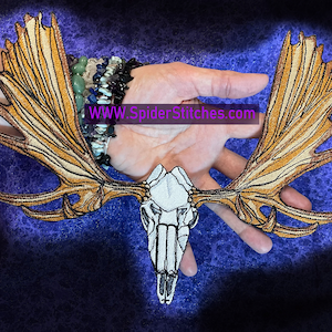 以下が含まれることがあります： 白い刺繍のヘラジカの頭蓋骨で、大きく詳細な角があります。角は薄い茶色で、質感があります。頭蓋骨は濃い青色の背景に囲まれています。