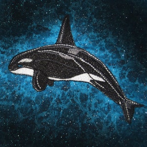 Può includere: Una silhouette di orca nera e bianca ricamata su uno sfondo di tessuto blu scintillante.