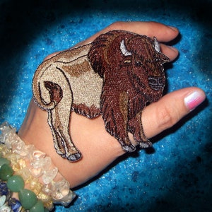 May include: A brown and tan embroidered patch of a bison with a white background. The patch is on a person's hand.