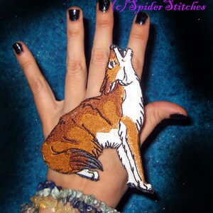 May include: A brown and white embroidered patch of a howling wolf, with a white background, is attached to a person's hand. The patch is a detailed, realistic depiction of a wolf with a white chest and a brown body. The wolf is howling with its mouth open and its ears perked up. The patch is attached to the person's hand with a small piece of fabric.