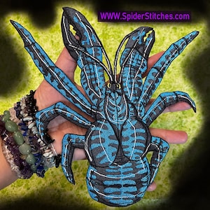 Op de afbeelding: Een blauw en zwart geborduurde spinnenpatch met witte accenten. De spin heeft grote klauwen en een gedetailleerd lichaam. De tekst "WWW.SpiderStitches.com" is zichtbaar. De patch wordt in een hand gehouden.