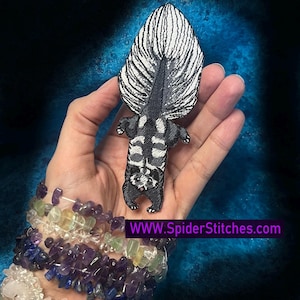 Könnte beinhalten: Gestickter Skunk-Aufnäher mit schwarz-weißem Design. Der Skunk wird von unten betrachtet und zeigt seinen gestreiften Schwanz und seine Körpermarkierungen. Der Aufnäher wird vor einem blauen Hintergrund gehalten. Die Website-Adresse ist sichtbar.