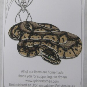 May include: Iron-on patch featuring a detailed embroidered snake with green eyes. The patch is on a white card with the text "Iron on Patch" and "www.spiderstitches.com".