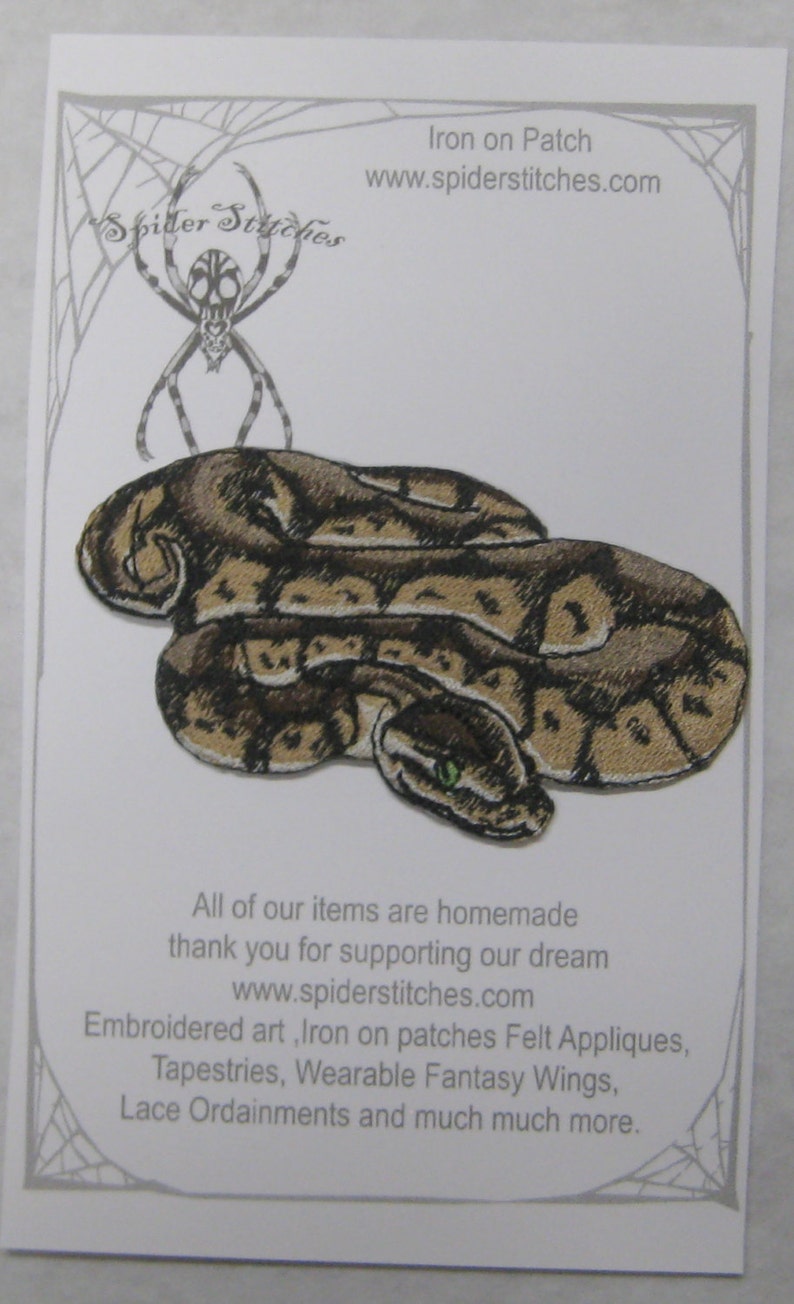 May include: Iron-on patch featuring a detailed embroidered snake with green eyes. The patch is on a white card with the text "Iron on Patch" and "www.spiderstitches.com".