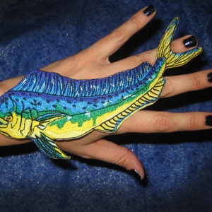 May include: A blue, yellow, and green embroidered patch of a mahi-mahi fish. The patch is on a person's hand.